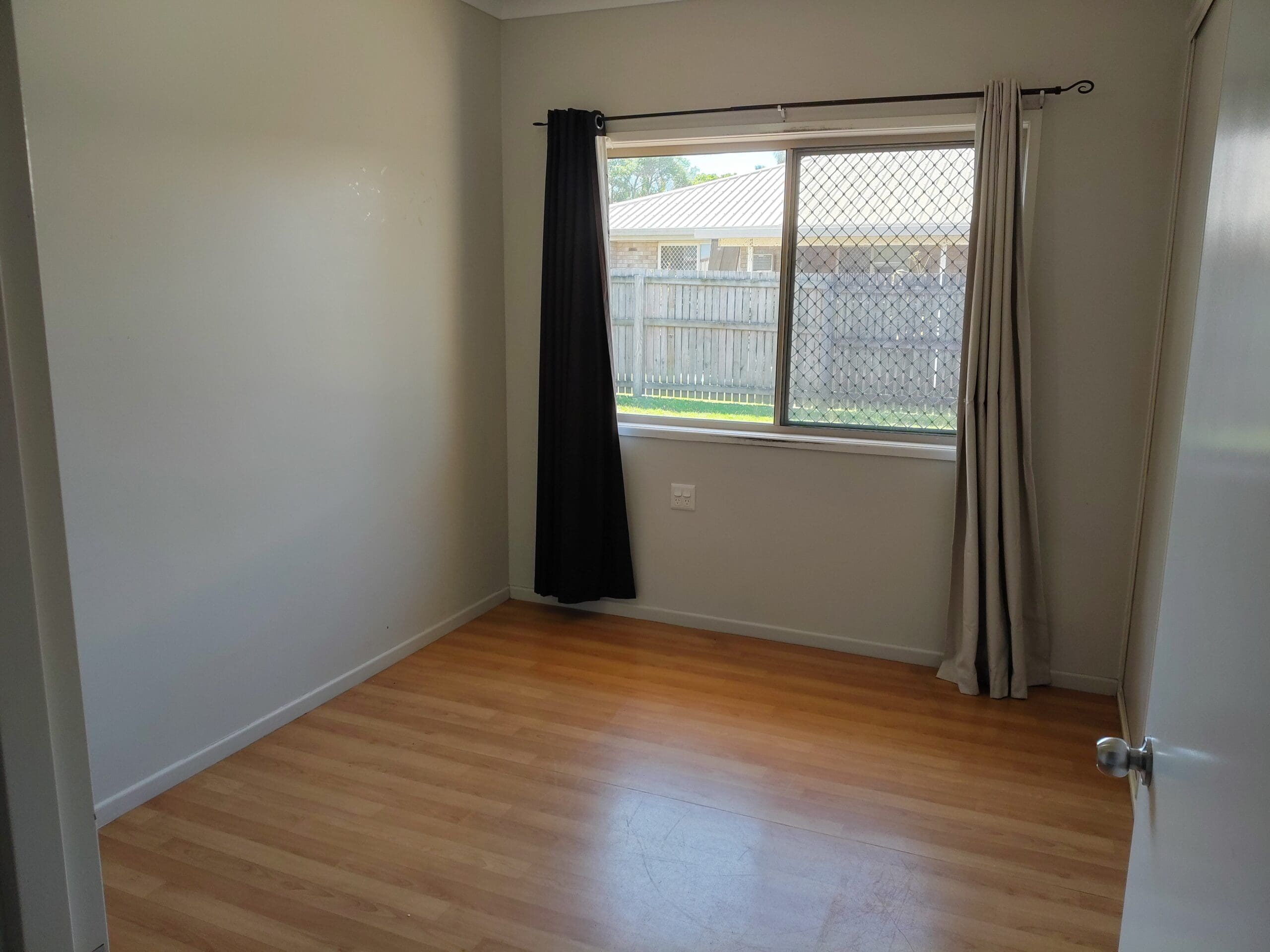 SIL Home in Mackay, Inverness Court Beaconsfield bedroom with timber-look flooring, curtained window and fenced yard view by Australia's leading disability accommodation provider.