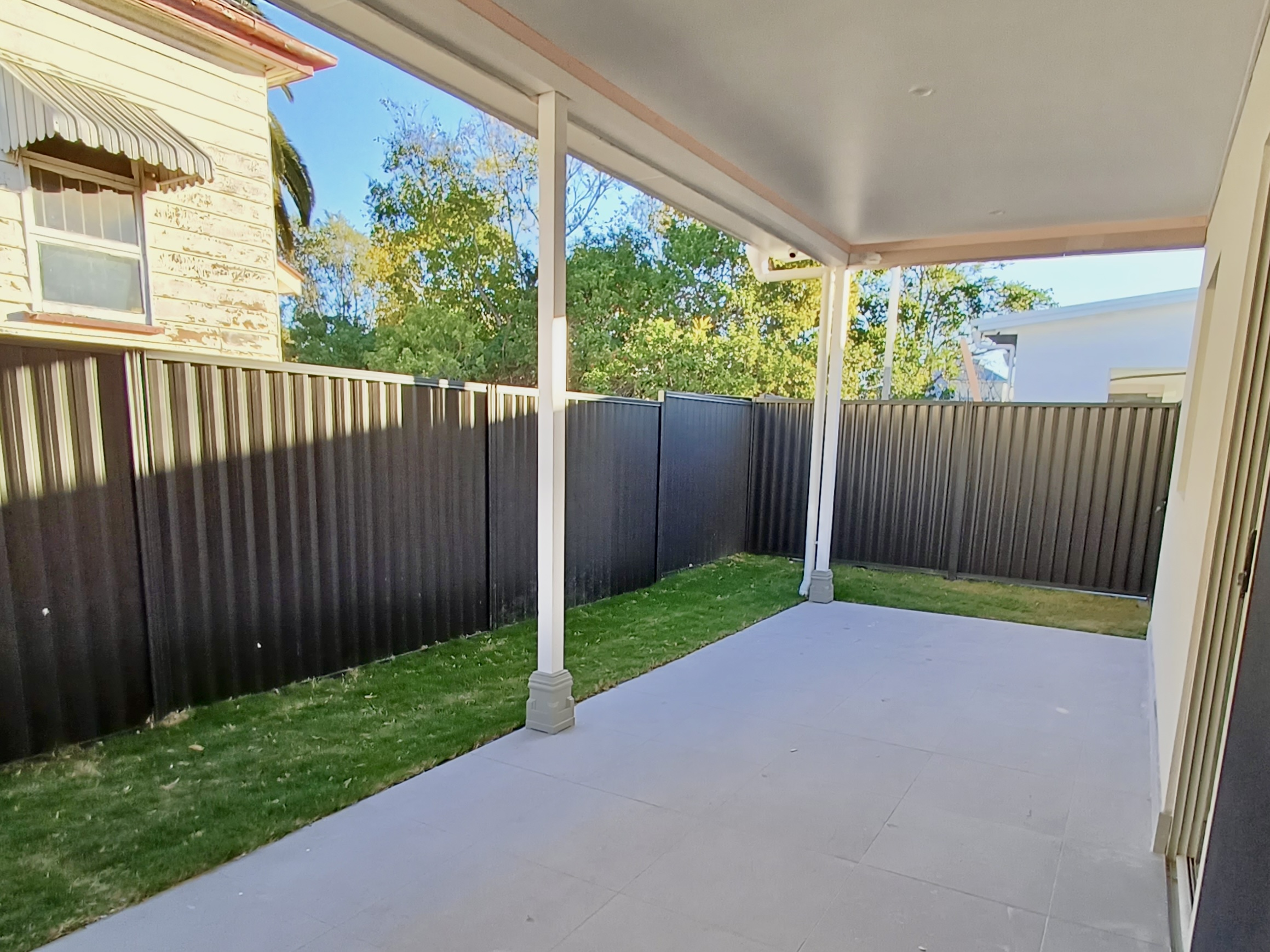 Covered alfresco area at Kanda Booval SDA Unit 1 featuring a concrete floor, small lawn area, and a high black corrugated metal fence for privacy.