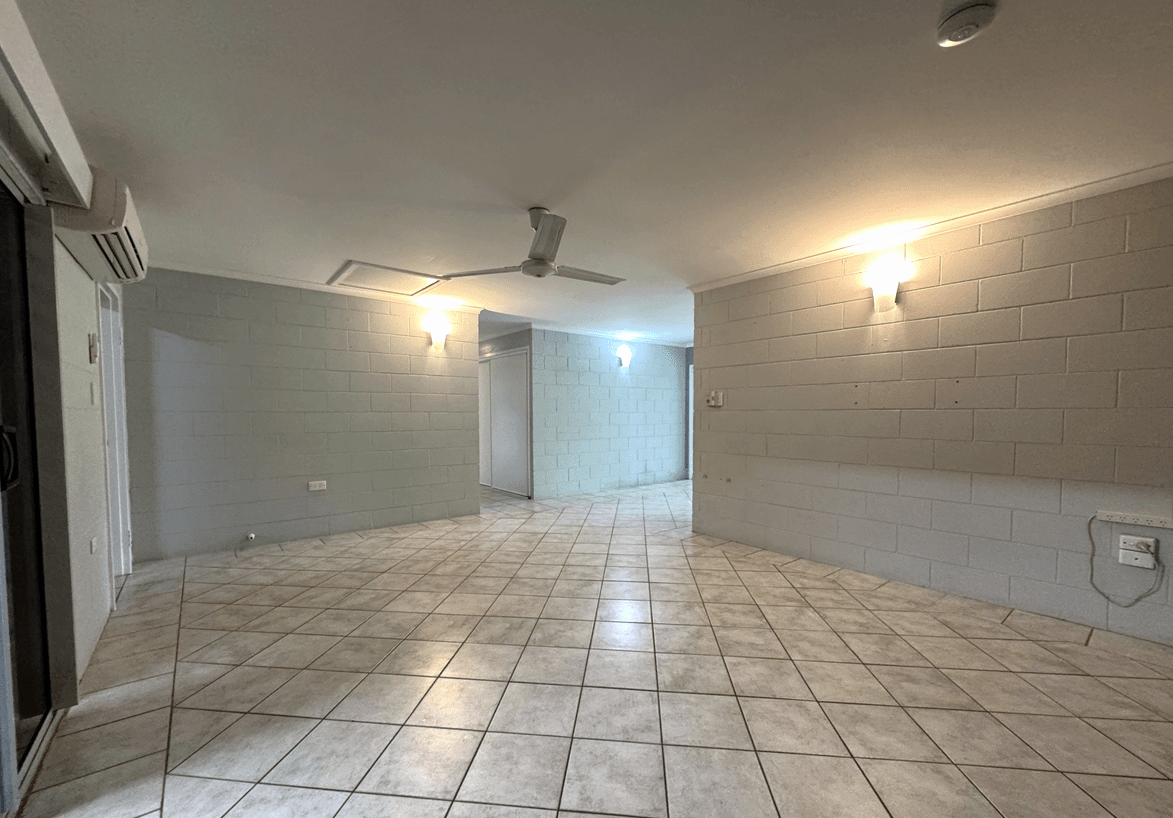 Spacious, unoccupied living area in the Edmonton Supported Independent Living (SIL) Accommodation. The room features large tiled flooring, light grey block-style walls, wall sconce lighting, a ceiling fan, and an air conditioning unit. A sliding door and internal doorways are visible, leading to other parts of the home.