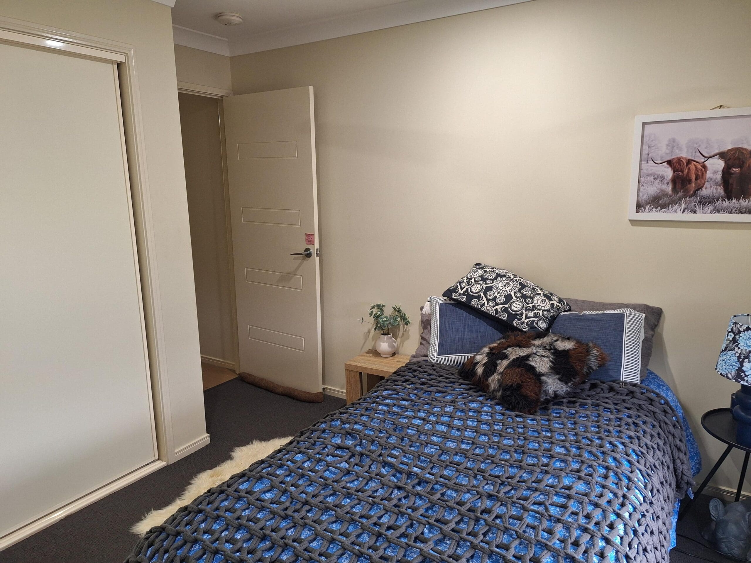 Bedroom in welcoming and cosy SIL in Brassall, Ipswich, featuring blue-grey bed, nightstand, and cow artwork, part of Australia’s leading disability accommodation provider.
